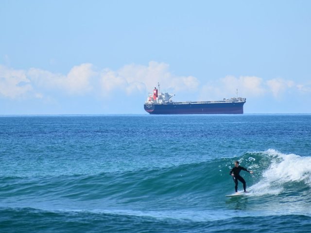 Surfer riding a wave with a bulk carrier ship on the horizon