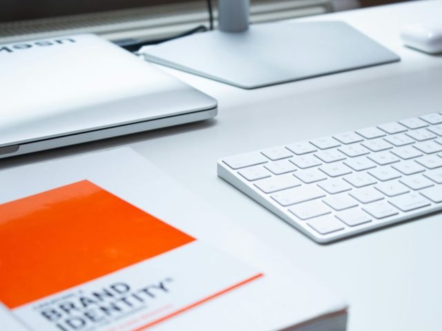 White desk with a laptop, keyboard and an orange brand identity book